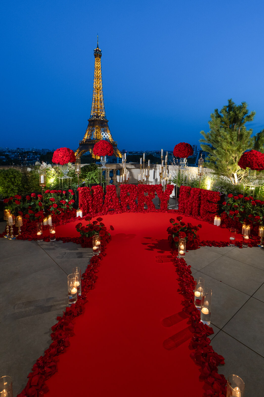 Romantic "Marry Me" letters proposal package in Paris with a luxurious rooftop setup at the Shangri-La Hotel. Features red letters, abundant red roses, endless candles, and the Eiffel Tower illuminated against the night sky.