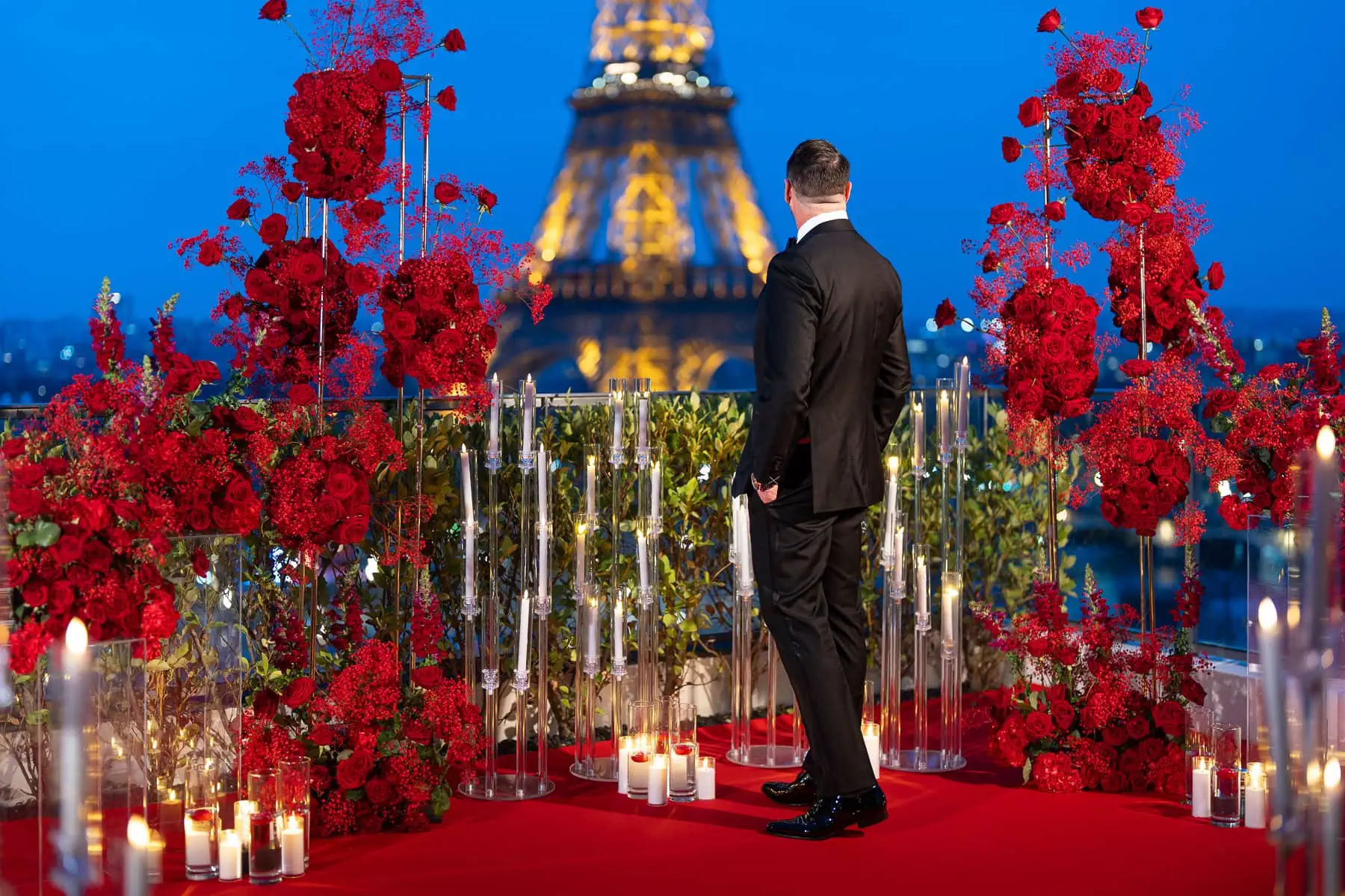 Paris Proposal Planner sets a luxurious scene on the Shangri-La Eiffel Tower terrace: a sumptuous red carpet, roses, and candles beneath a vivid blue sky, with a gentleman awaiting his partner.