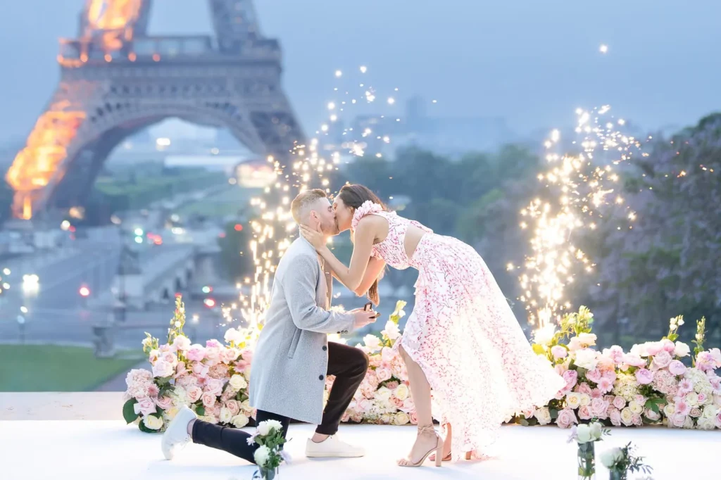 Man proposing at sunrise in front of the Eiffel Tower with pink floral arrangements and fireworks at Trocadéro, Paris.