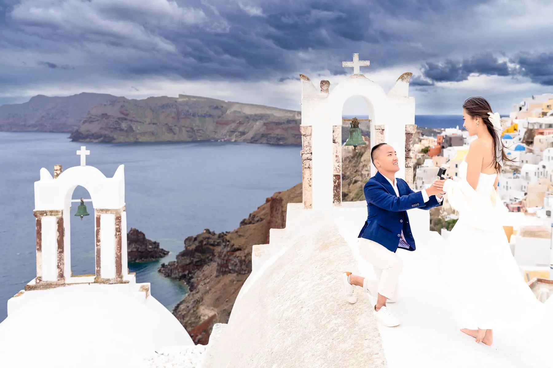 Man proposing on one knee with ring box to woman on exclusive Santorini rooftop with iconic blue domes and colorful houses in background - Destination proposal planning by Kiss Me in Paris.